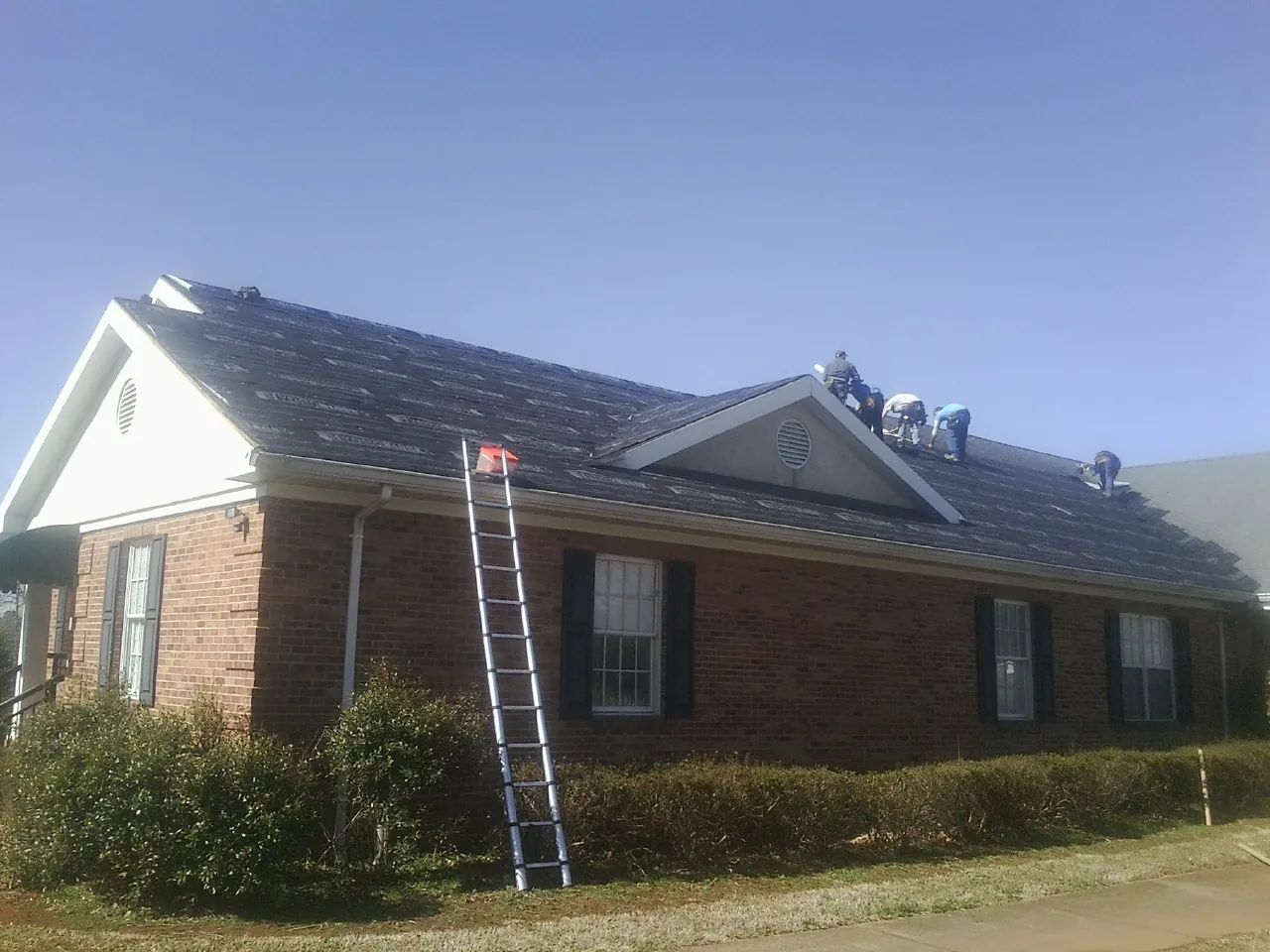 Crew tearing shingles off a brick ranch roof with a ladder set — Emergency Roof Repair work by our crew in Altavista