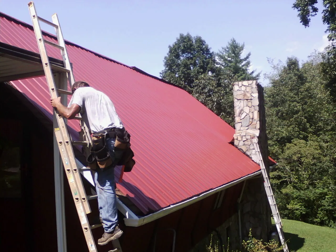 Roofer on a ladder beside a red metal roof and stone chimney — Soffit & Fascia Repair by Roofing Brothers in Altavista